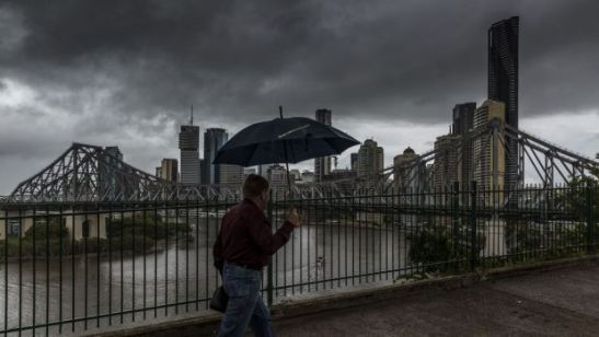 Painting of a man, with an open umbrella, walking along a riverside promenade on a rainy day. A bridge and skyscrapers in background.